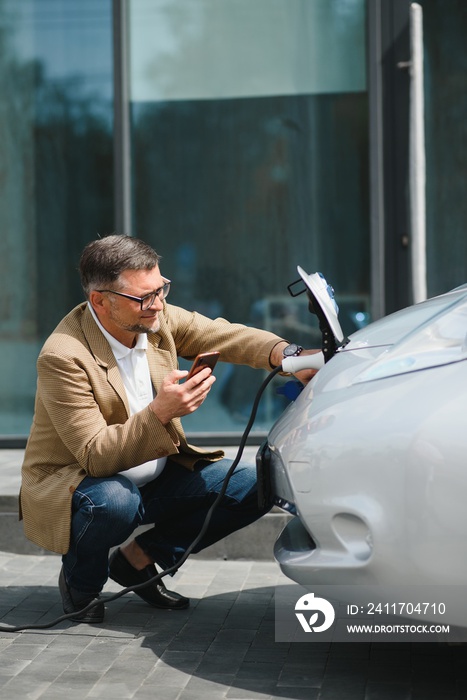 Hansome bearded man sitting near his new modern electric car and holding plug of the charger, while car is charging at the charging station