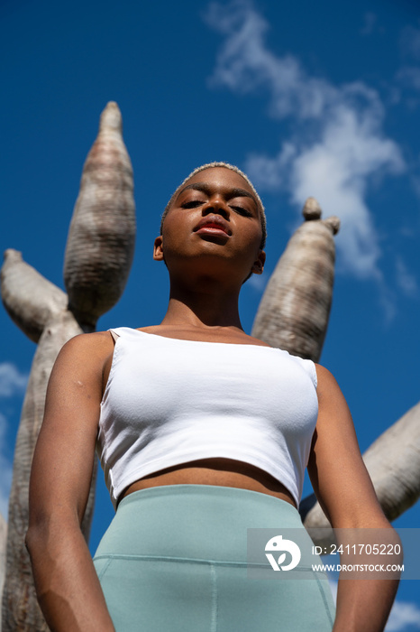 Confident African American female athlete in activewear and with short hair standing against dragon tree in park and looking at camera