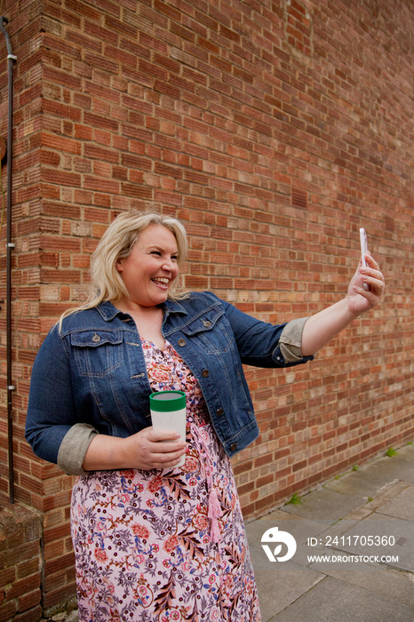 Plus-sized woman in front of brick wall taking a selfie with a reusable coffee cup