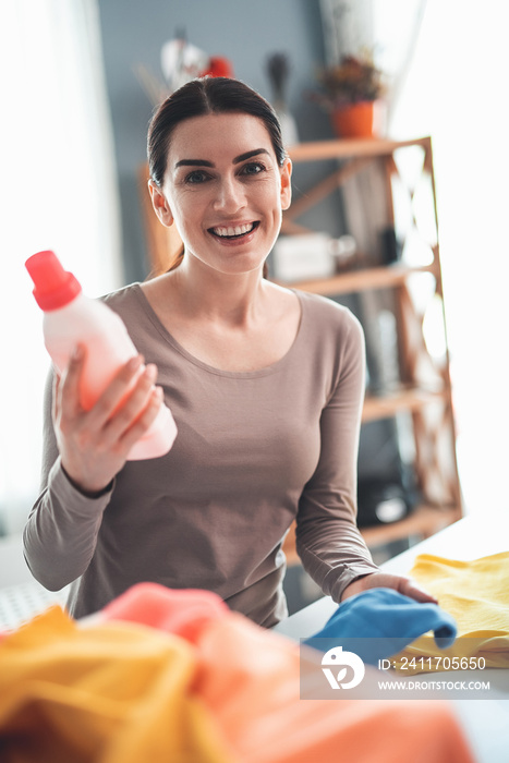 Waist up portrait of content woman holding pink detergent with happy face. She satisfied with using washing liquid to remove stains from dirty clothes