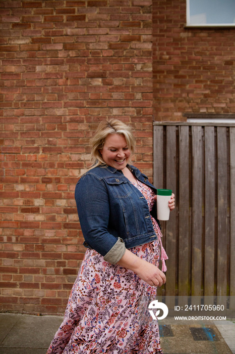 Plus-sized woman in front of brick wall holding a reusable coffee cup and smiling