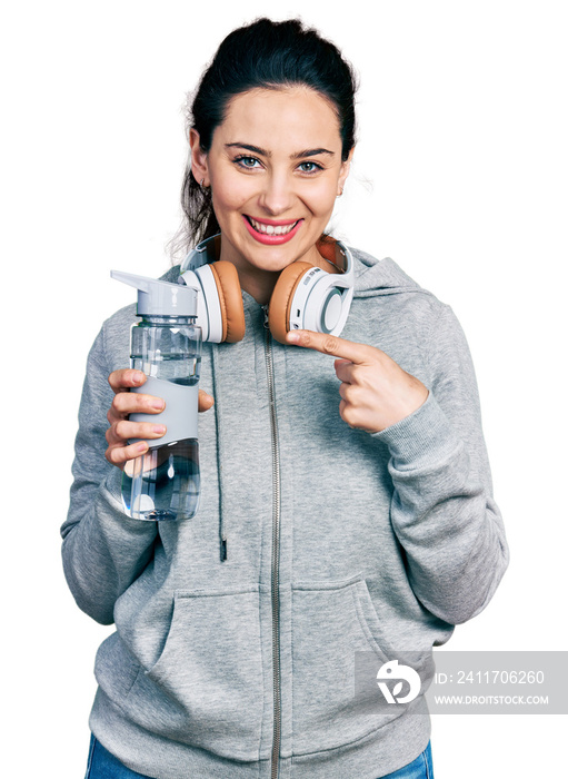 Young hispanic woman wearing sportswear holding water bottle using headphones smiling happy pointing with hand and finger