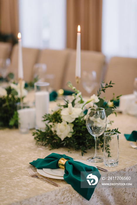 Wedding set up, dinner table reception. A plate with a green cloth towel, knives and forks next to the plate. Flower composition with eucalyptus leaves in the center of the table and burning candles.