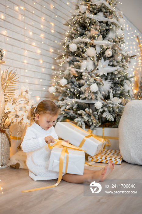 Very nice charming little girl blonde in white dress sitting near Christmas trees in bright interior of the house