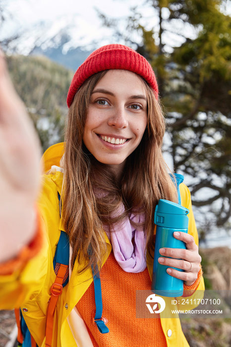 Beautiful smiling woman holds unrecognizable device, makes selfie, wears red hat and yellow jacket, holds blue flask, drinks hot beverage, models against beautiful landscape. Picturesque view