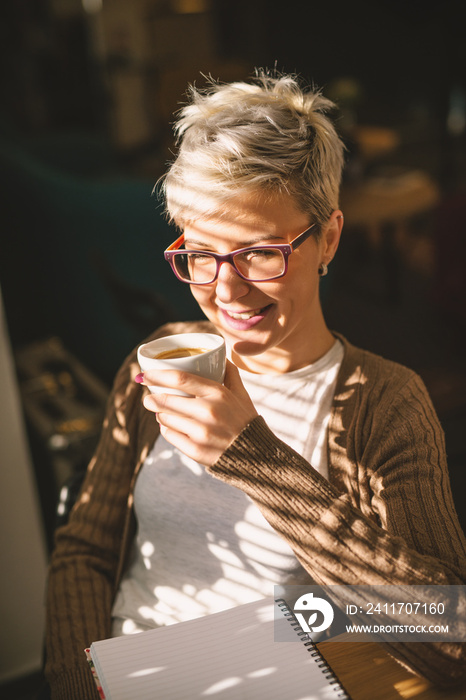 Portrait of beautiful smiling blond female drinking coffee while sitting near the window with a diary on the table.