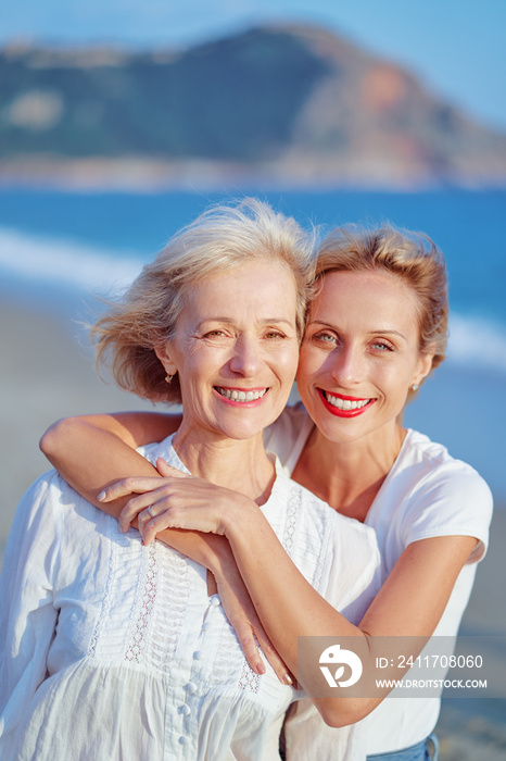 Outdoor close up portrait of smiling happy caucasian senior mother with her adult daughter hugging and looking at the camera on sea beach.