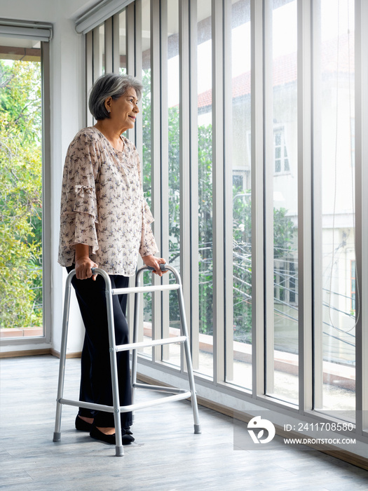 Asian senior woman, white hair standing with walking frame, full length, looking out the glass window, vertical style. Elderly lady patient using walking frame. Strong health, medical care concepts.