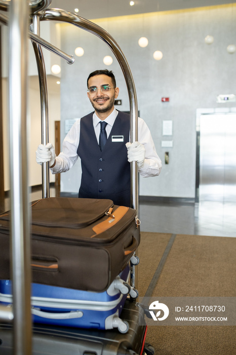 Young hotel porter in uniform pushing cart with baggage of travelers