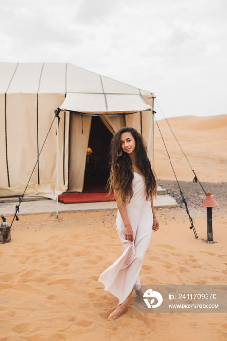 Portrait of beautiful oriental young brunette woman in white dress. Glamping tent in moroccan desert. Luxury modern camp in Morocco.