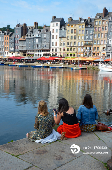 The old port (Vieux Bassin) of the town Honfleur in the county of Calvados in Normandy, France, three young ladies on the quay in front, blurred norman houses in the background, sunny day in summer