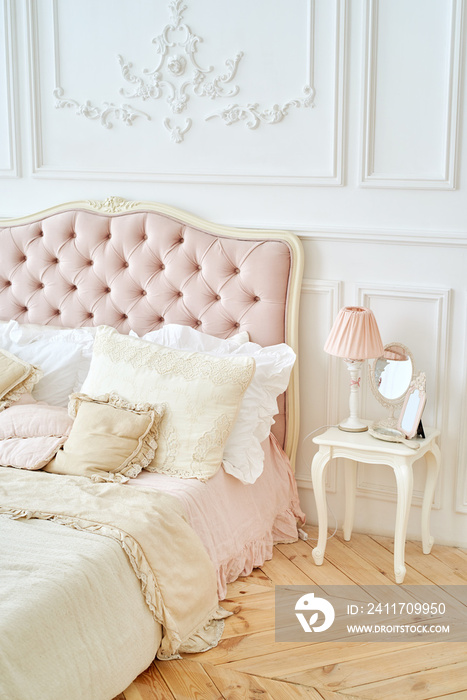 Elegant home or hotel bedroom interior. Pillows on luxury buttoned bed and  pink lamp, mirror on white wooden table, vintage style