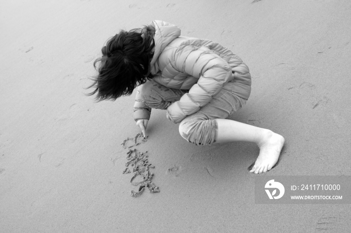 Ein Junge mit langen Haaren und Daunenjacke schreibt etwas in den Sand am Strand in Knokke-Heist an der Nordsee bei Brügge in Westflandern in Belgien, fotografiert in klassischem Schwarzweiß