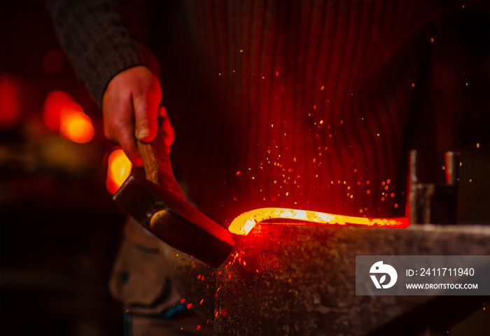 The blacksmith hits the red-hot workpiece in the forge with a hammer and glowing sparks fly in all directions