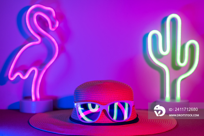 Summer hat with sunglasses refection flamingo and cactus neon pink and blue and green light on table with copy space.Trendy vacation holiday background.