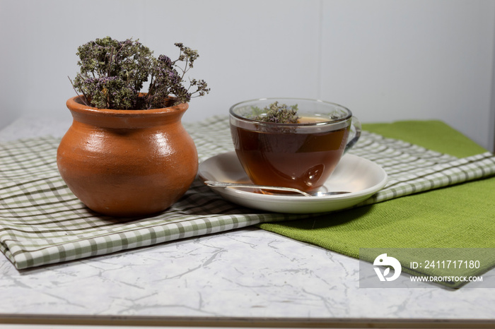 A cup of tea with a floating oregano flower stands on a white saucer, on which lies a silver spoon. Next to the cup is a clay pot with a bouquet of oregano.