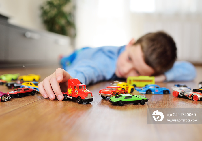 a preschool boy in a blue sweater is lying on the wooden floor playing with toy cars against the background of a bright room