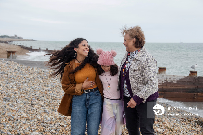 Grandmother, mother and daughter laughing while walking on beach on cloudy day