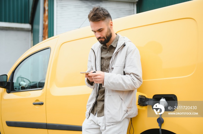 bearded caucasian man standing near an electric car that is charging and making time adjustments on a smartphone.