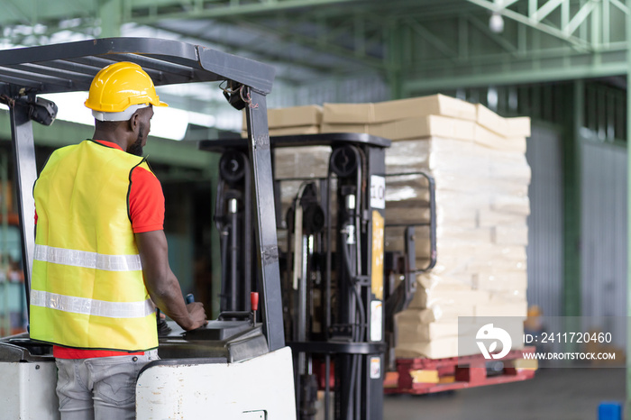 African American male warehouse worker in safety vest and helmet driving and operating on forklift truck for transfer products or parcel goods in the industrial storage warehouse. multicultural worker