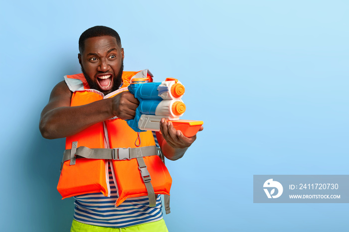 Waterfight battle. Emotional black man screams I will shoot you, holds toy water gun, has fun during summer vacation with friends, plays game at beach, dressed in lifevest, stands against blue wall