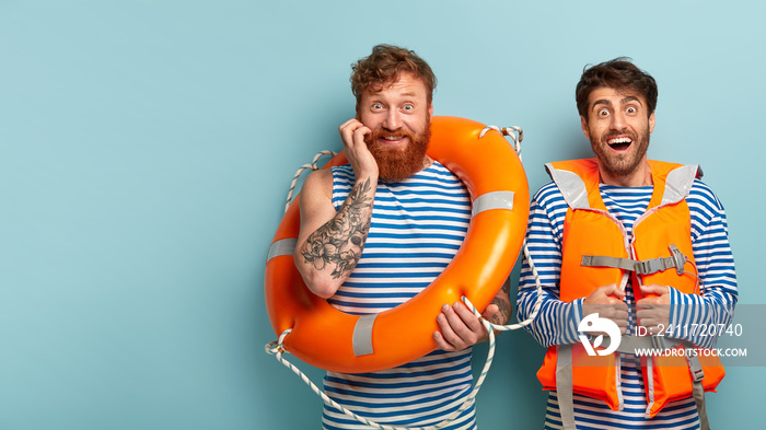 Happy successful lifesavers look gladly at camera, watch situation at sea, enjoy nature, pose with orange lifering and inflatable lifejacket, pose against blue background, free space on left. Summer