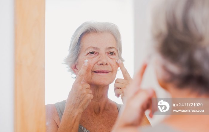 Portrait of senior beautiful gray-haired woman applies anti aging cream on wrinkled face looking at the mirror - take care of the skin concept