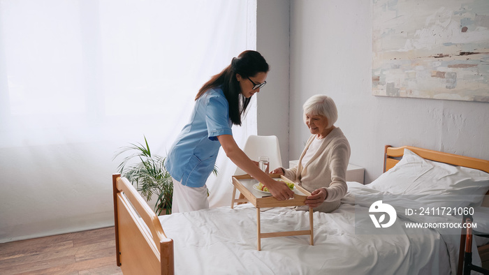 brunette nurse carrying tray with breakfast to smiling aged patient