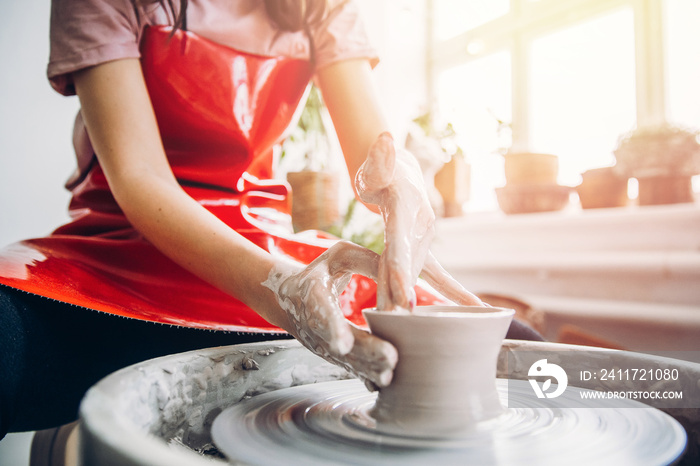 Young woman in red apron works behind potter wheel with length, making handmade plate. Concept of concentration, creativity hand