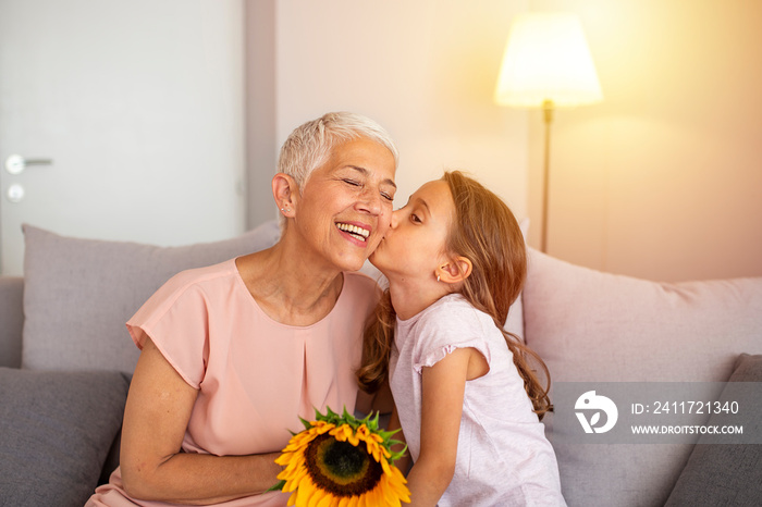 Little preschool granddaughter kissing happy older grandma on cheek giving violet flowers bouquet congratulating smiling senior grandmother with birthday, celebrating mothers day or 8 march concept