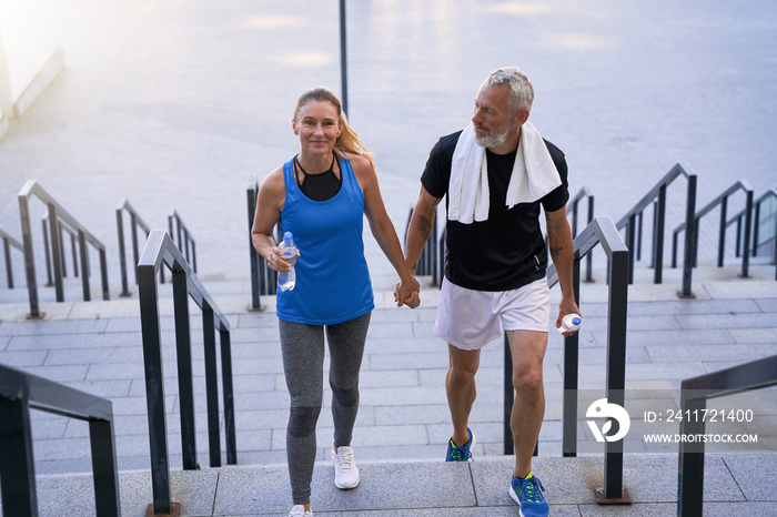 Sporty middle aged man and woman in sportswear looking happy, holding hands while walking up the stairs after training together in the city