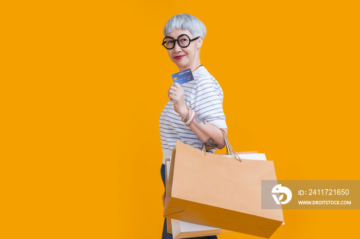 Asian happy senior female woman tattoo holds colourful shopping packages standing on yellow background studio shot, Close up Portrait old female hand hold shopping bags enjoy shopping with joyful