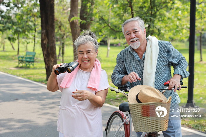 Portrait of happy asian senior man and woman walking and hugging with bicycle and binoculars in summer garden outdoor. Lover couple going to picnic at the park. Happiness marriage lifestyle concept.