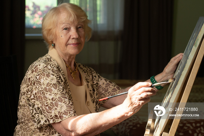 An elderly gray-haired woman sits at the easel and paints in oils, sitting against the window in the sunlight. Silver age concept.