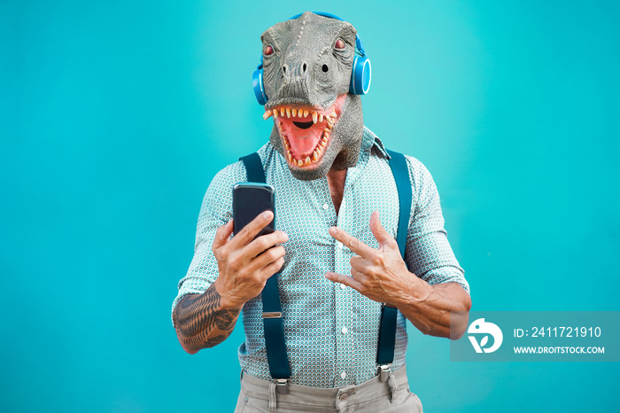 Tattooed man with t-rex mask using smartphone while listening music - Crazy senior guy choosing playlist from mobile phone app - Technology trends and madness costume concept - Focus on face