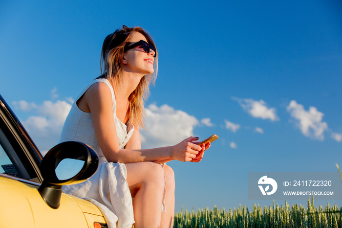 young girl in sunglasses with smartphone is sitting on a car on green wheat field in spring time