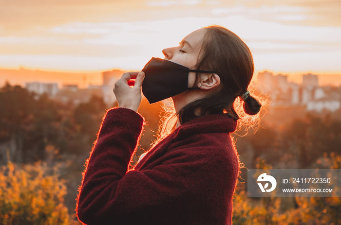 Side view of young woman taking the face mask off for a gulp of fresh air backlit by orange sunset light