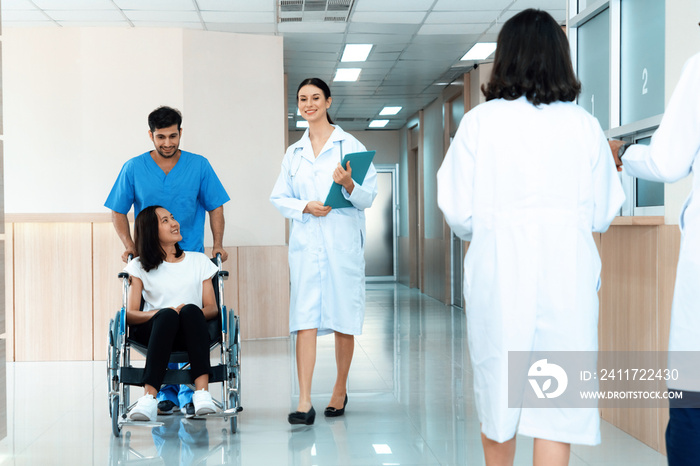 Doctor and male nurse transport a female patient in a wheelchair along sterile hospital corridor. Health care and nursing care for disabled handicapped patient in the hospital concept.