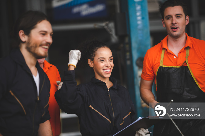 Group of young and senior male and female car mechanics in garage wearing uniform enjoying and celebrating with female worker holding clipping board