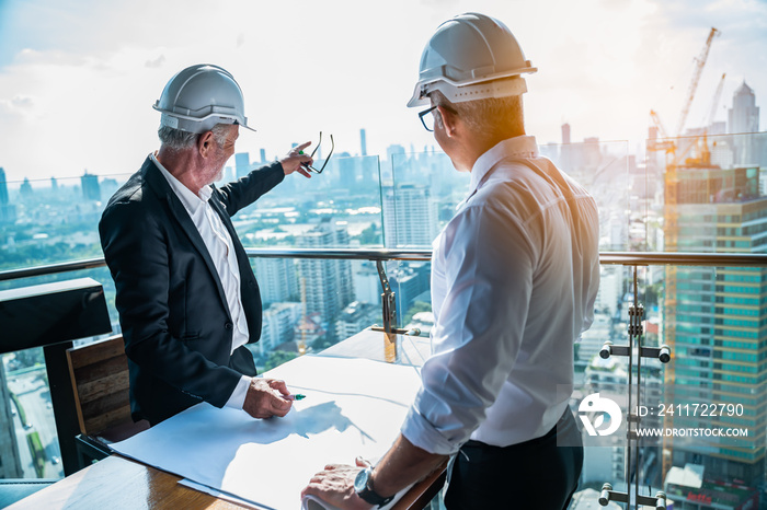 Two Engineer or Architect are analyzing blueprints while working on a new project on construction site with blue sky and city background.Architect supervising construction on terrace tower.