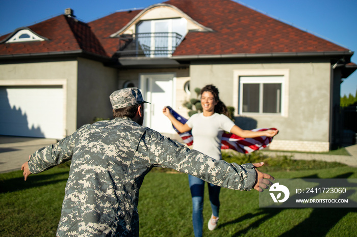 Soldier in uniform coming home and his lovely wife with American flag running into his arms celebrating reunion.