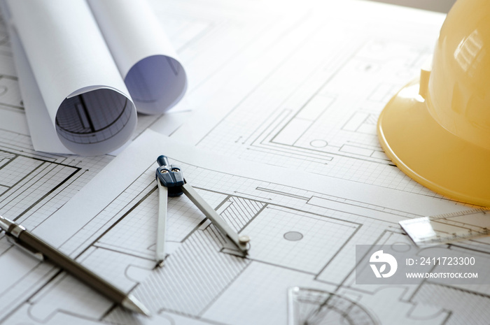 Yellow hard safety helmet hat and architectural blueprints and blueprint rolls and pencils pen, ruler, protractor, and tape measure on the table at the construction site.