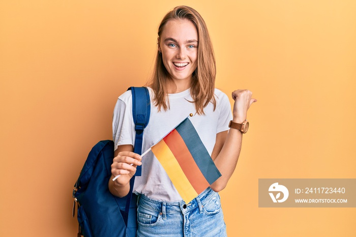 Beautiful blonde woman exchange student holding germany flag pointing thumb up to the side smiling happy with open mouth