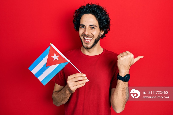Handsome hispanic man holding cuba flag pointing thumb up to the side smiling happy with open mouth