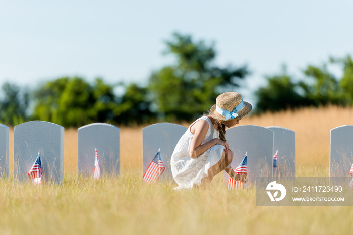 selective focus of kid in dress and straw hat sitting near headstone with american flag in graveyard