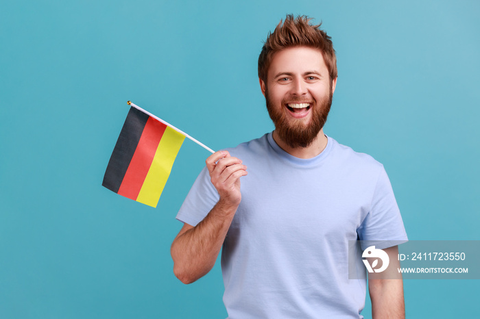 Portrait of delighted satisfied bearded man holding Germany flag, celebrating Day of Germany - 3th October, expressing positive emotions. Indoor studio shot isolated on blue background.