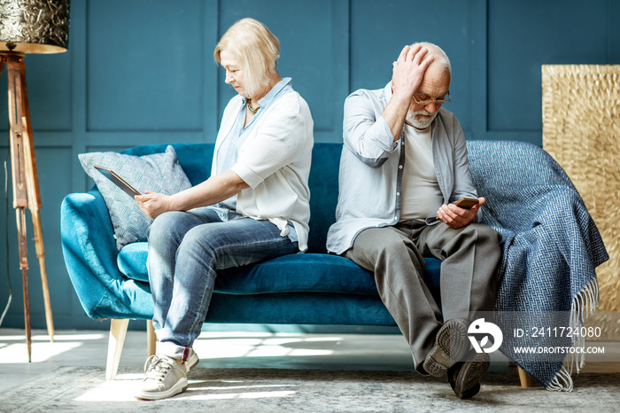 Offended senior man and woman sitting back to each other, using digital gadgets on the couch at home