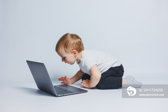 Little smart boy 3-4 years old sits with a laptop on a white background. A child in a white T-shirt and black trousers sits at a laptop and looks at the screen. Modern progressive children