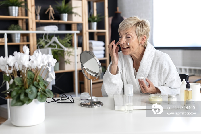 An older woman with short blonde hair in a white bathrobe sits at the table and puts cream on her face inside the living room