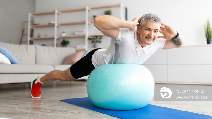 Happy senior man doing exercises with fitness ball at home, working out his back muscles, training with sports equipment